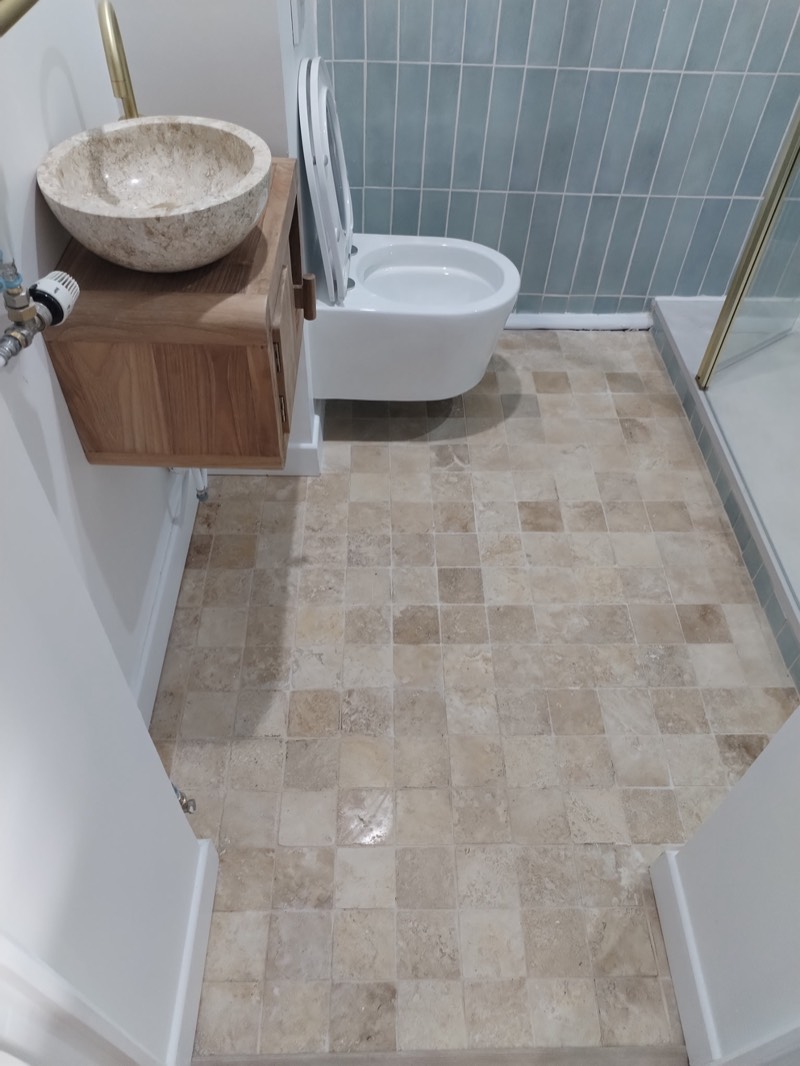 Après : salle d'eau rénovée — travertin au sol, faïence bleu clair, WC suspendu, vasque pierre, robinetterie dorée